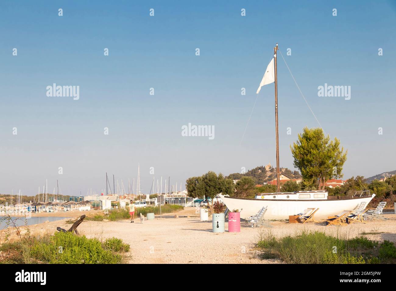 Tribunj, Croacia - 16 de agosto de 2021: Hombre caminando un perro en la playa vacía por el café bar con un barco a vela en la orilla Foto de stock