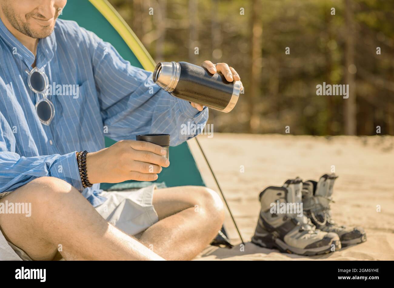 Sonriente hombre caucásico está sentado al lado de su tienda de turismo y sosteniendo una botella de té termo en su mano al aire libre. Foto de stock