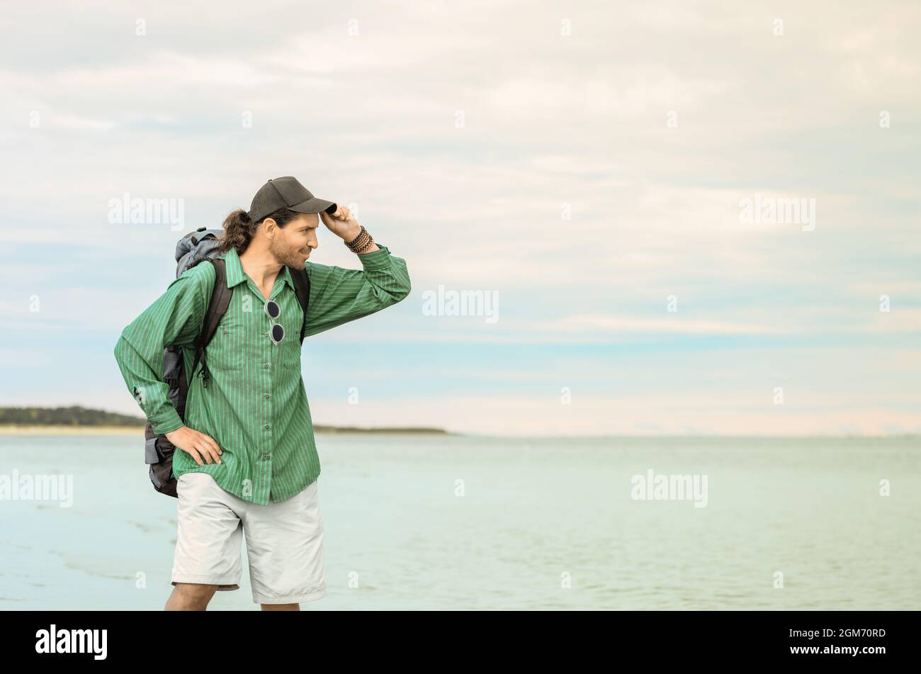 El viajero con su mochila turística está de pie sobre un fondo marino. El hombre está quitándose su gorra de béisbol y mirando a una distancia. Foto de stock