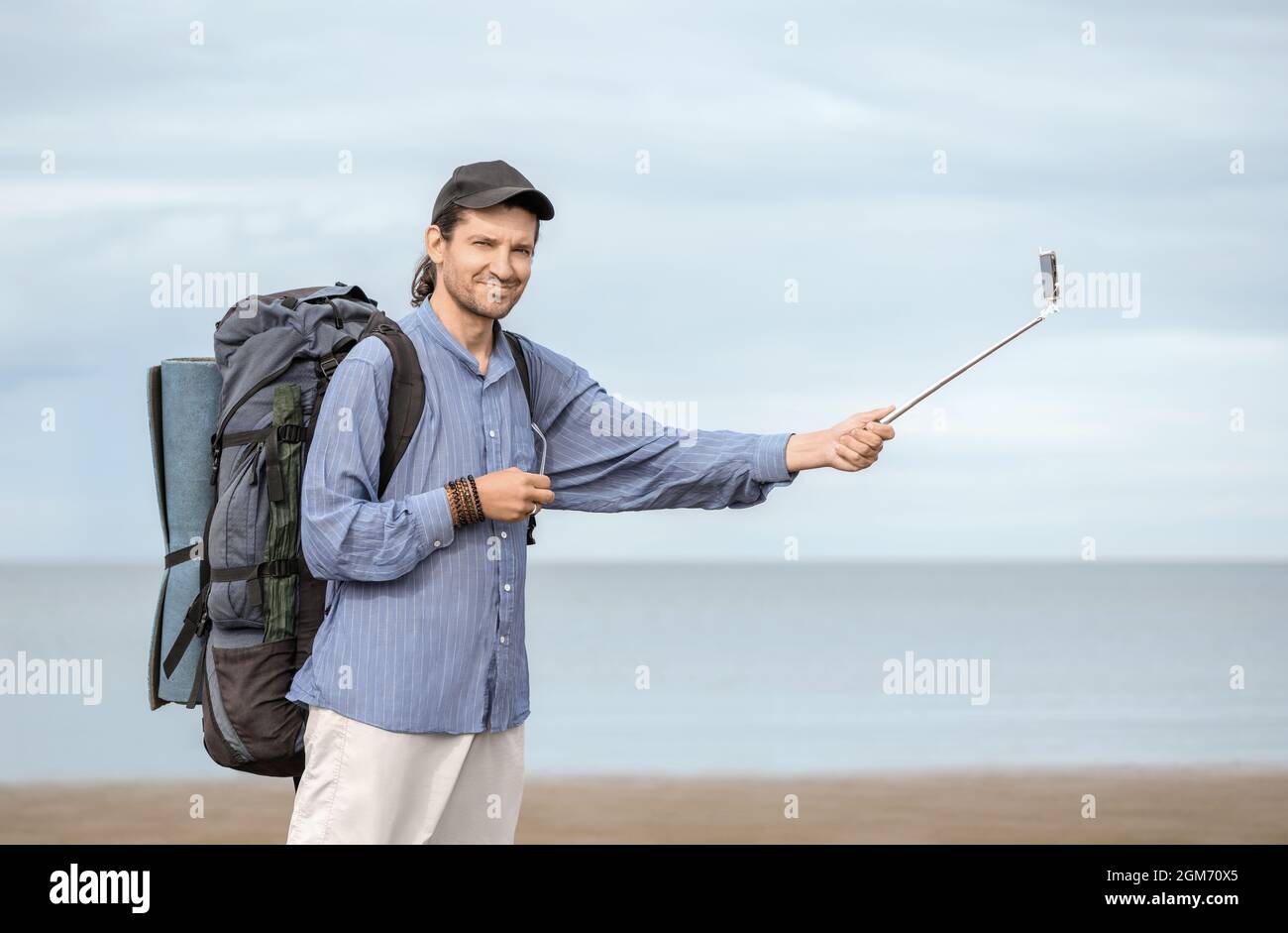 Un hombre de 40 años de edad, con su mochila y estera turística sostiene un palo de selfie blanco en su mano contra el fondo de un mar en un día nublado. Foto de stock