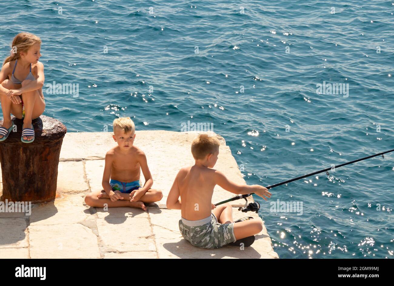 Prvic Sepurine, Croacia - 25 de agosto de 2021: Niños rubios sentados al sol en el muelle de piedra pescando y mirando la distancia, y un mar detrás de t Foto de stock