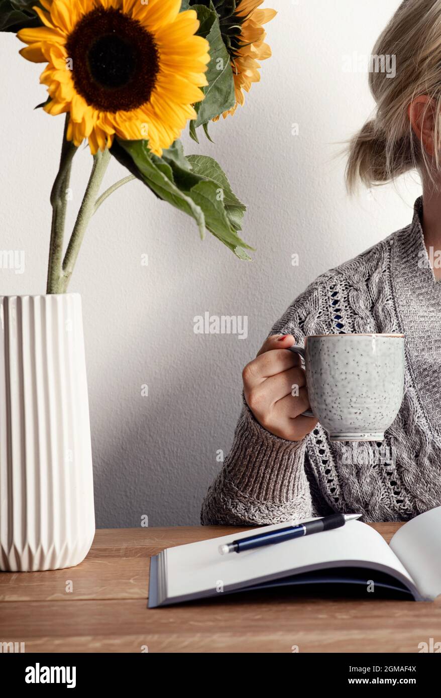 Mujer con taza de café en su mano mientras está sentada en su oficina con  cuaderno, bolígrafo y jarrón con girasoles frescos en la mesa de madera  Fotografía de stock - Alamy