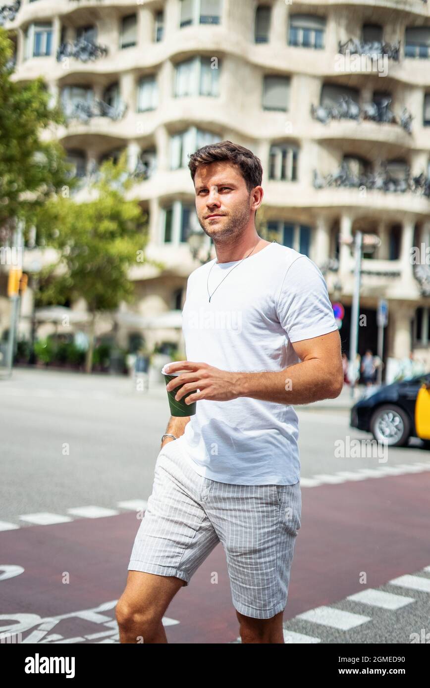 Hombre joven con barba en ropa de verano informal y moderna con una taza de  bebida para llevar caminando por la carretera pavimentada cerca del  edificio en un día soleado en la