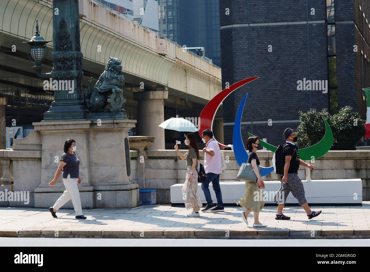 La gente pasa por un monumento de símbolo paralímpico junto a una estatua de un león en el puente de Nihonbashi durante los Paralímpicos de Tokio en un día caluroso. Foto de stock