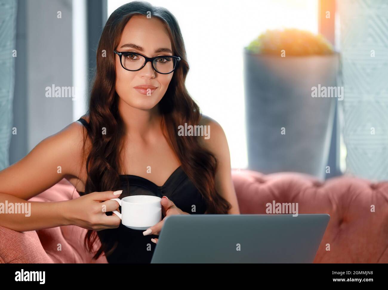 Una hermosa joven europea en vasos de 20-30 años se sienta en el sofá y sostiene una taza de café o té. Una chica hermosa trabaja remotamente en un ordenador portátil. Foto de stock