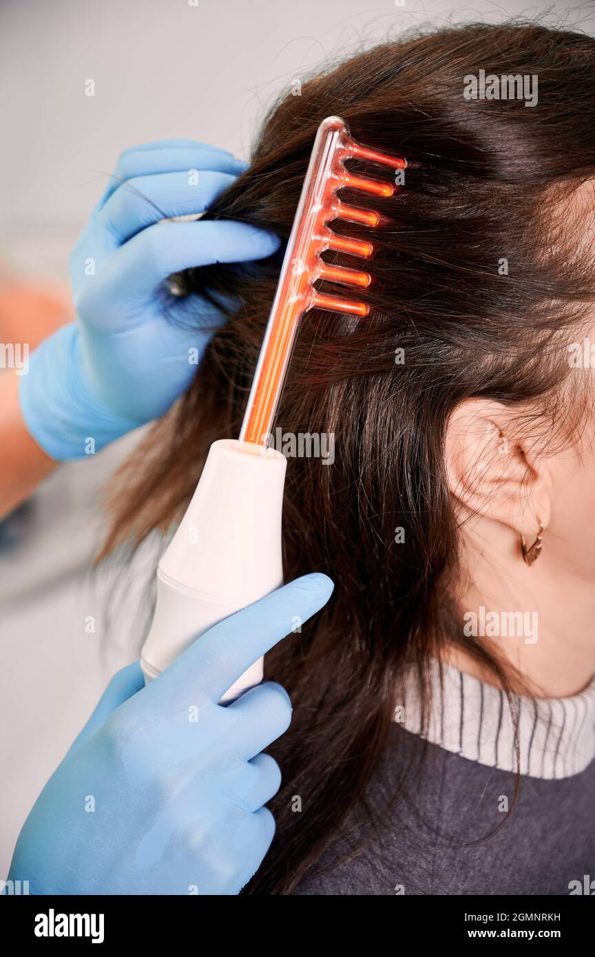 Primer plano de la mano del médico trichologist en guantes estériles  cepillando el pelo femenino del cliente con el dispositivo del crecimiento  del pelo del laser. Mujer que recibe tratamiento del cabello