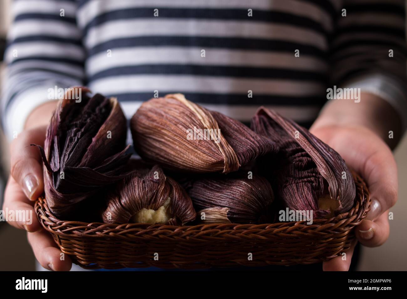 Persona que sostiene muchos alimentos tradicionales latinos hechos de maíz y envueltos en hojas de maíz púrpura en una cesta sobre una mesa de madera negra Foto de stock