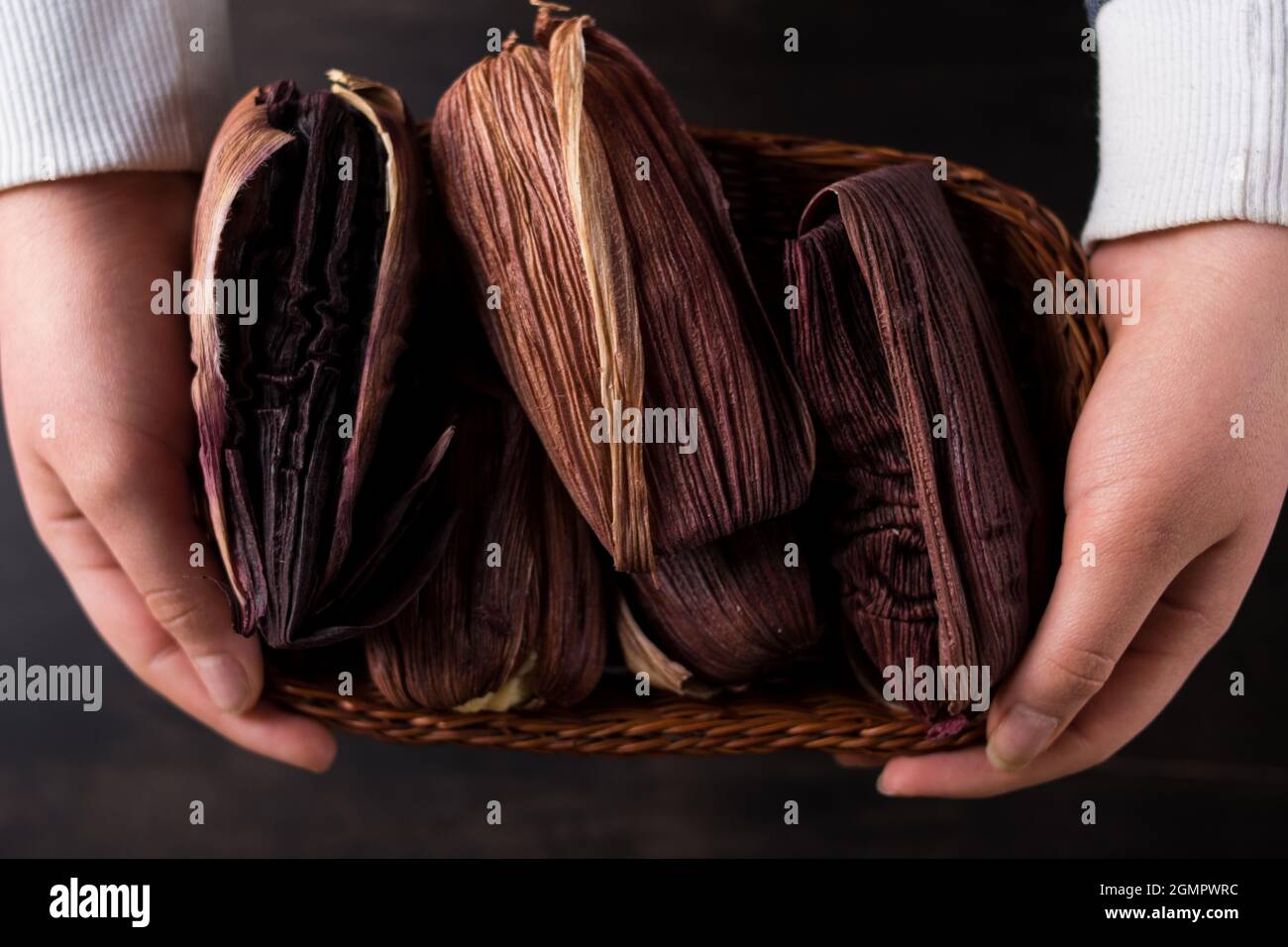 Persona que sostiene muchos alimentos tradicionales latinos hechos de maíz y envueltos en hojas de maíz púrpura en una cesta sobre una mesa de madera negra Foto de stock