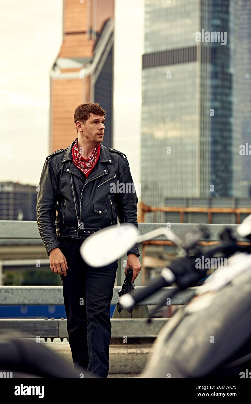 Retrato de un hombre elegante vestido con una chaqueta de cuero negro y  bandana sentado en su moto retro hecha a medida, mirando hacia fuera, los  edificios de la grande Fotografía de