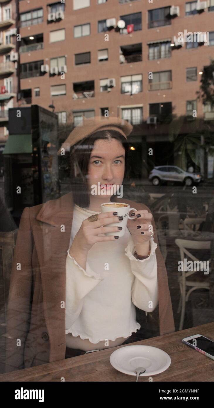 Mujer joven en la cafetería sosteniendo en sus manos una taza caliente de café listo para beber posando para la cámara. Estilo Vibes parisino para la temporada de otoño de 2021. Foto de stock