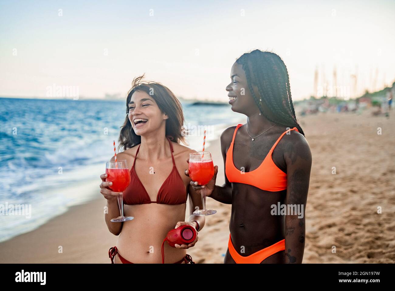 Jóvenes y diversas amigas en el bikini riendo felizmente mientras beben  cócteles rojos y escuchan música con altavoz de sonido portátil en la playa  de arena durante las vacaciones de verano juntos