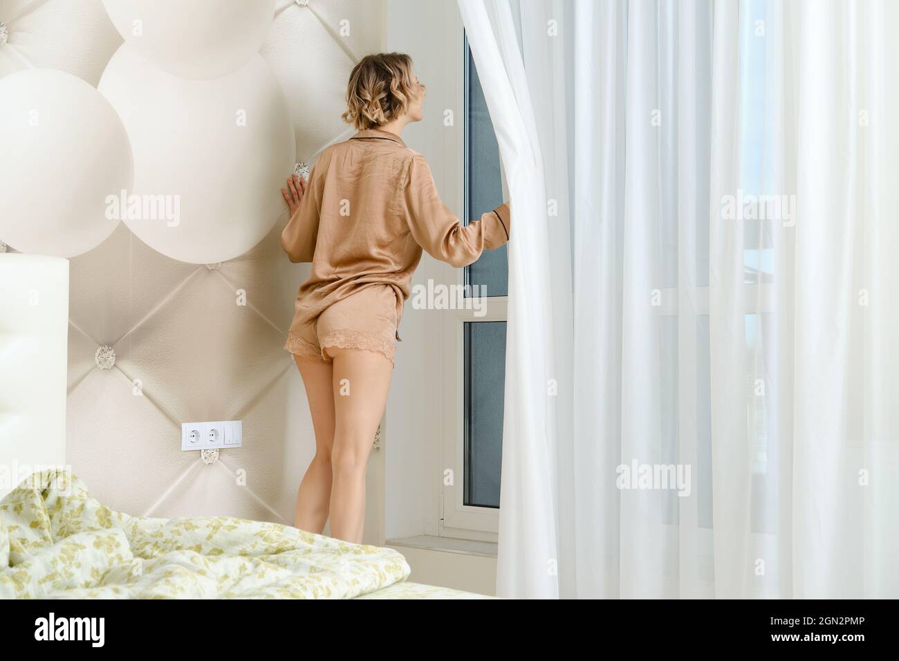 Una joven en pijama sale de la cama, llega a la ventana y mueve la cortina a un lado Foto de stock