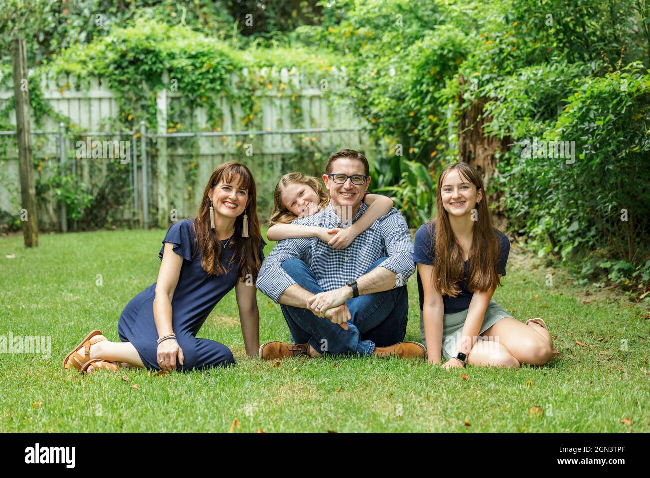 Una familia de una madre y un padre y dos hijas sentadas afuera en la hierba de su patio trasero con la hija más joven abrazando a papá de detrás Foto de stock