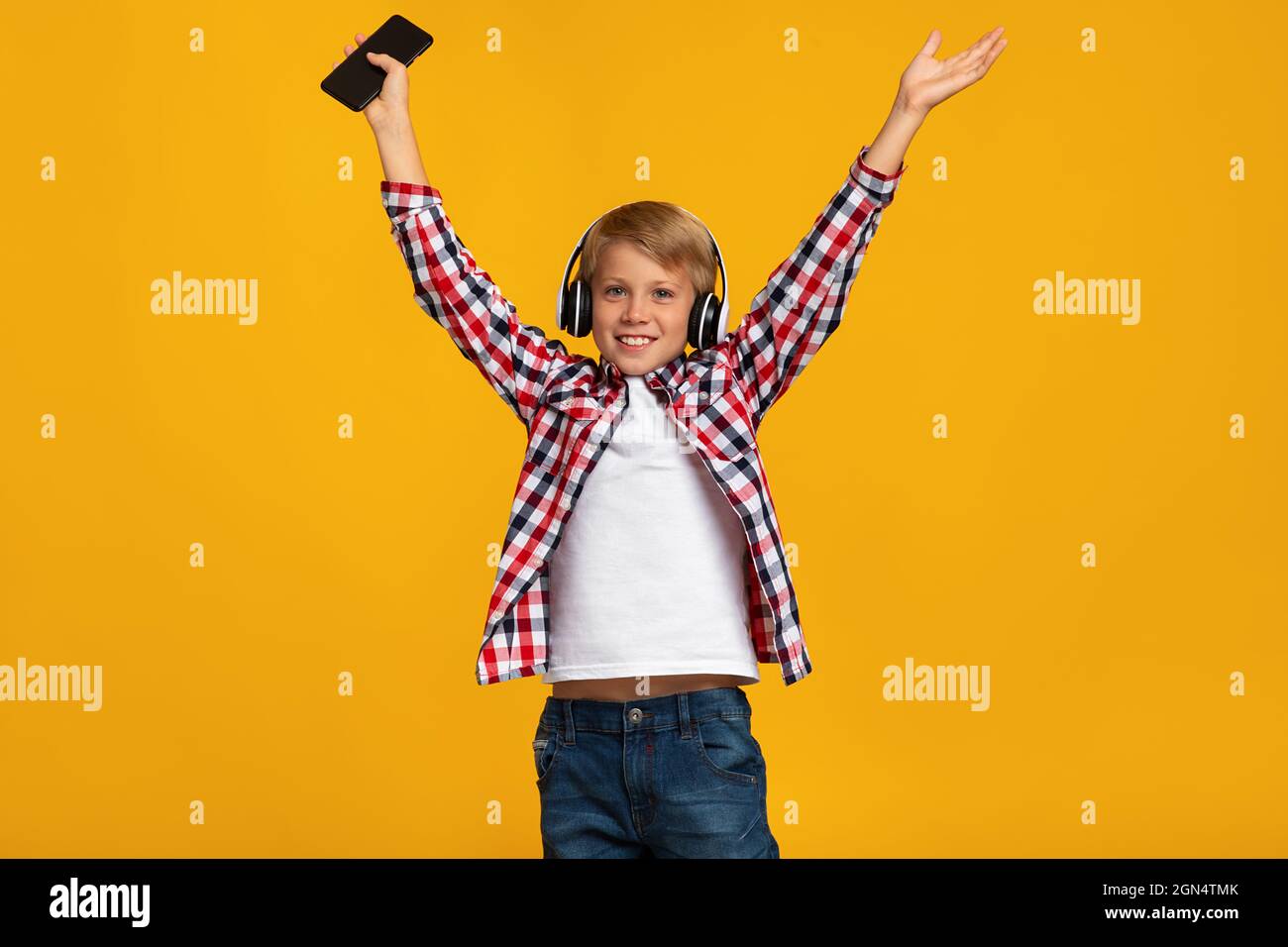 Feliz emoción feliz emocionado adolescente caucásico en auriculares sosteniendo smartphone, saltando, bailando y levantando las manos Foto de stock