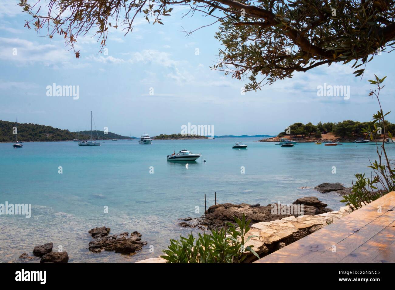 Kosirina, Murter, Croacia - 24 de agosto de 2021: Barcos amarrados en una bahía tranquila, vista desde el bar de la playa a través de la vegetación Foto de stock
