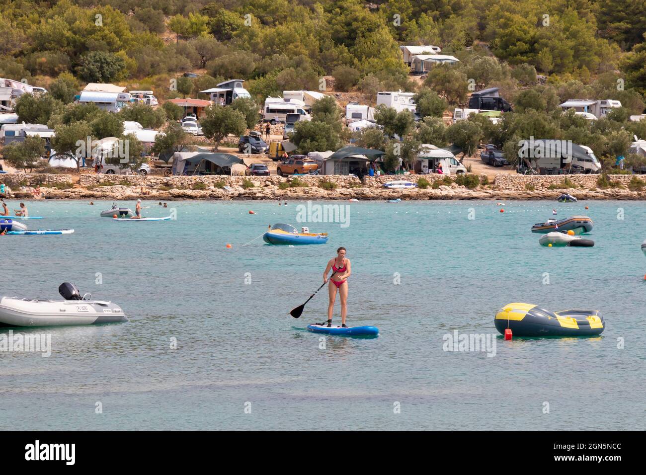 Kosirina, Murter, Croacia - 24 de agosto de 2021: Botes inflables amarrados en una tranquila bahía y gente remando en tablas de pie, en una playa de camping Foto de stock