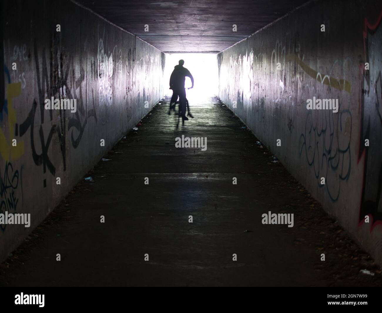 Con movimiento desenfocado y fuerte contraluz, dos jóvenes en un ciclo salen de un metro urbano a la luz del día con paredes cubiertas de graffiti a la izquierda y la derecha. Foto de stock