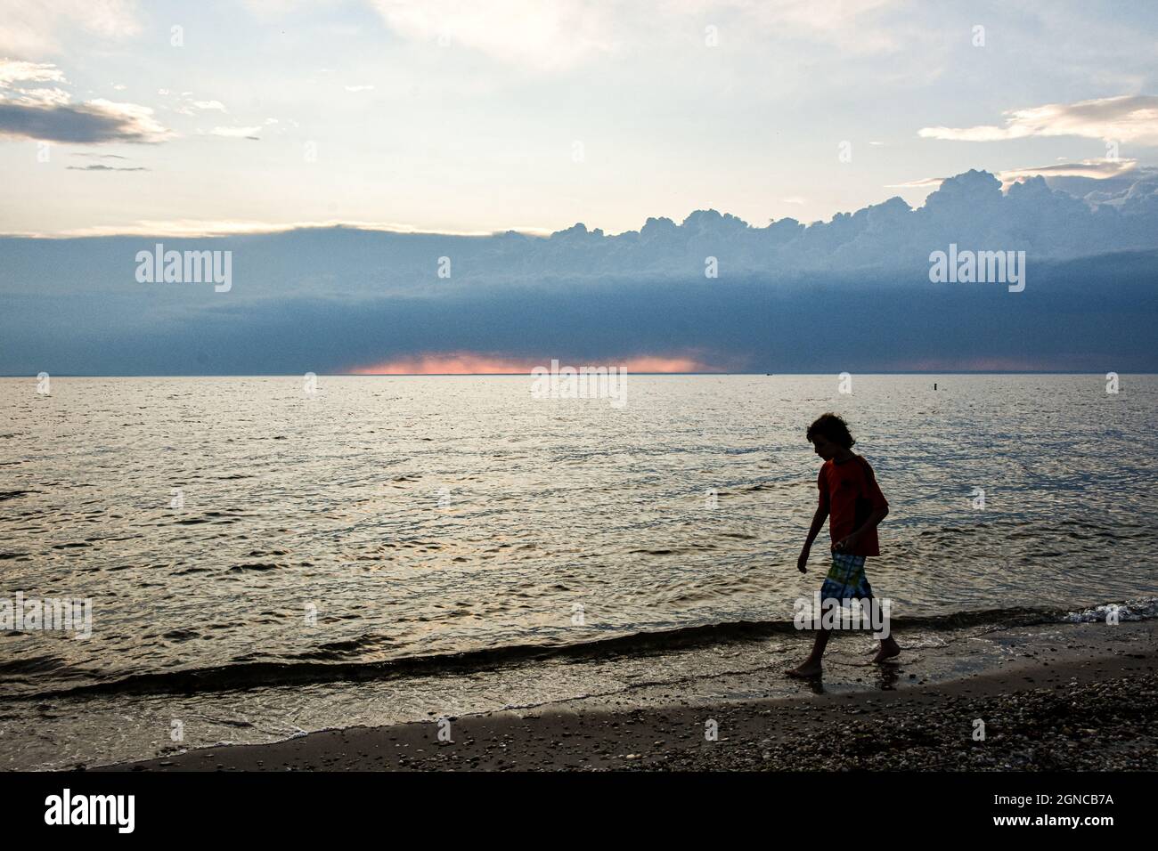 Un niño en silueta camina a lo largo de la playa con nubes de tormenta construyendo en el horizonte distante. Espacio de copia. Foto de stock