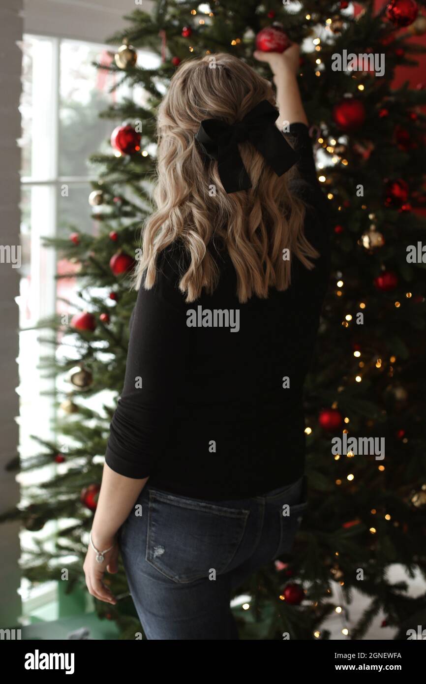 Hermosa mujer joven con modish peinado en un cómodo suéter y jeans que  adornan el árbol de Navidad en el interior. Mujer joven posando desde atrás  cerca Fotografía de stock - Alamy
