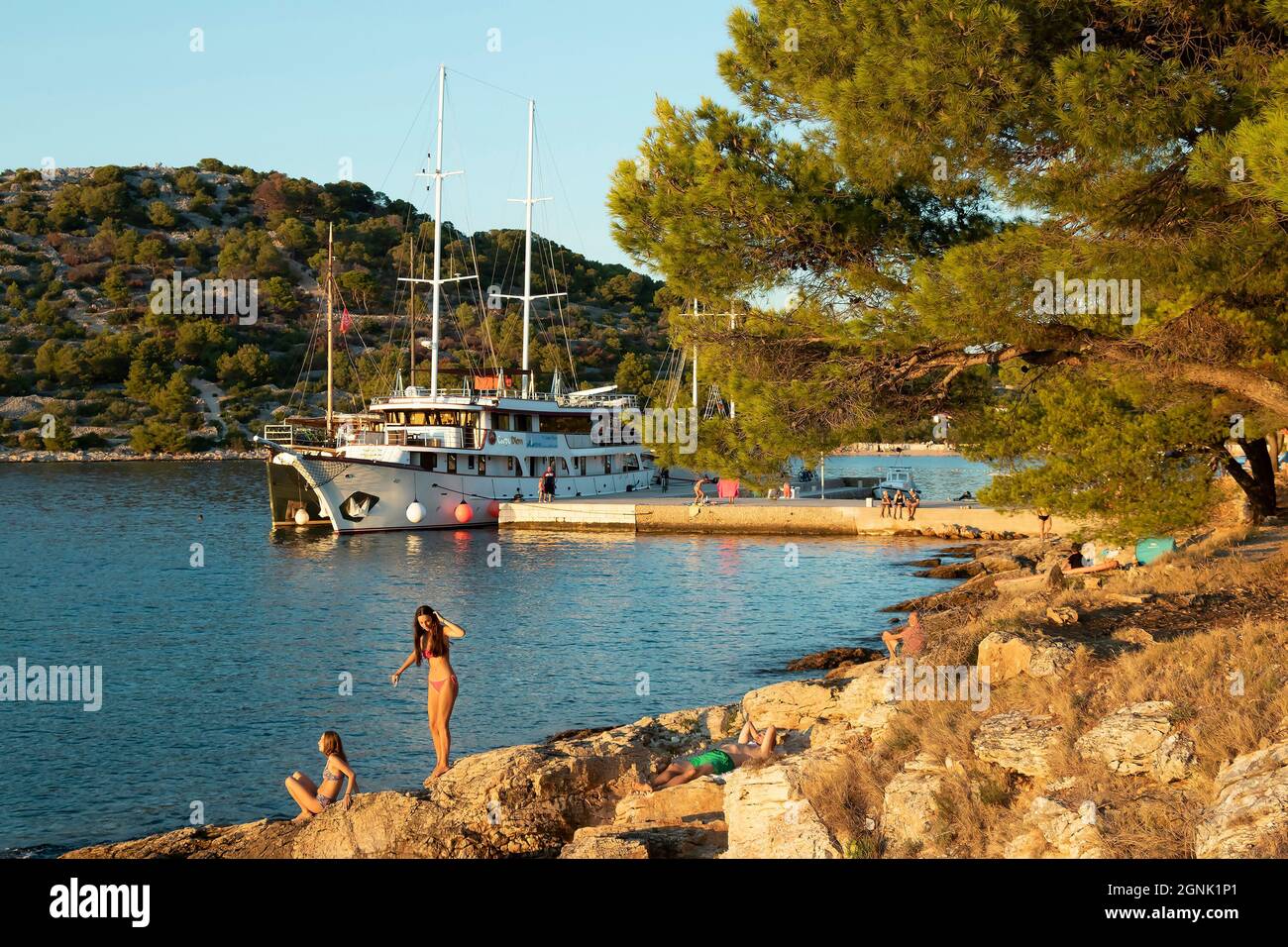 Podvrske, Murter, Croacia - 26 de agosto de 2021: Playa rocosa con pinos y personas en trajes de baño, y el barco atracó en el muelle detrás de ellos, en verano s. Foto de stock