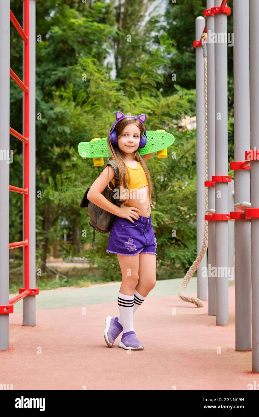 Retrato completo de la escuela primaria niña en ropa deportiva de moda y  auriculares inalámbricos en terreno deportivo. Niña posando con monopatín  durante las vacaciones de verano. Entretenimiento al aire libre Fotografía