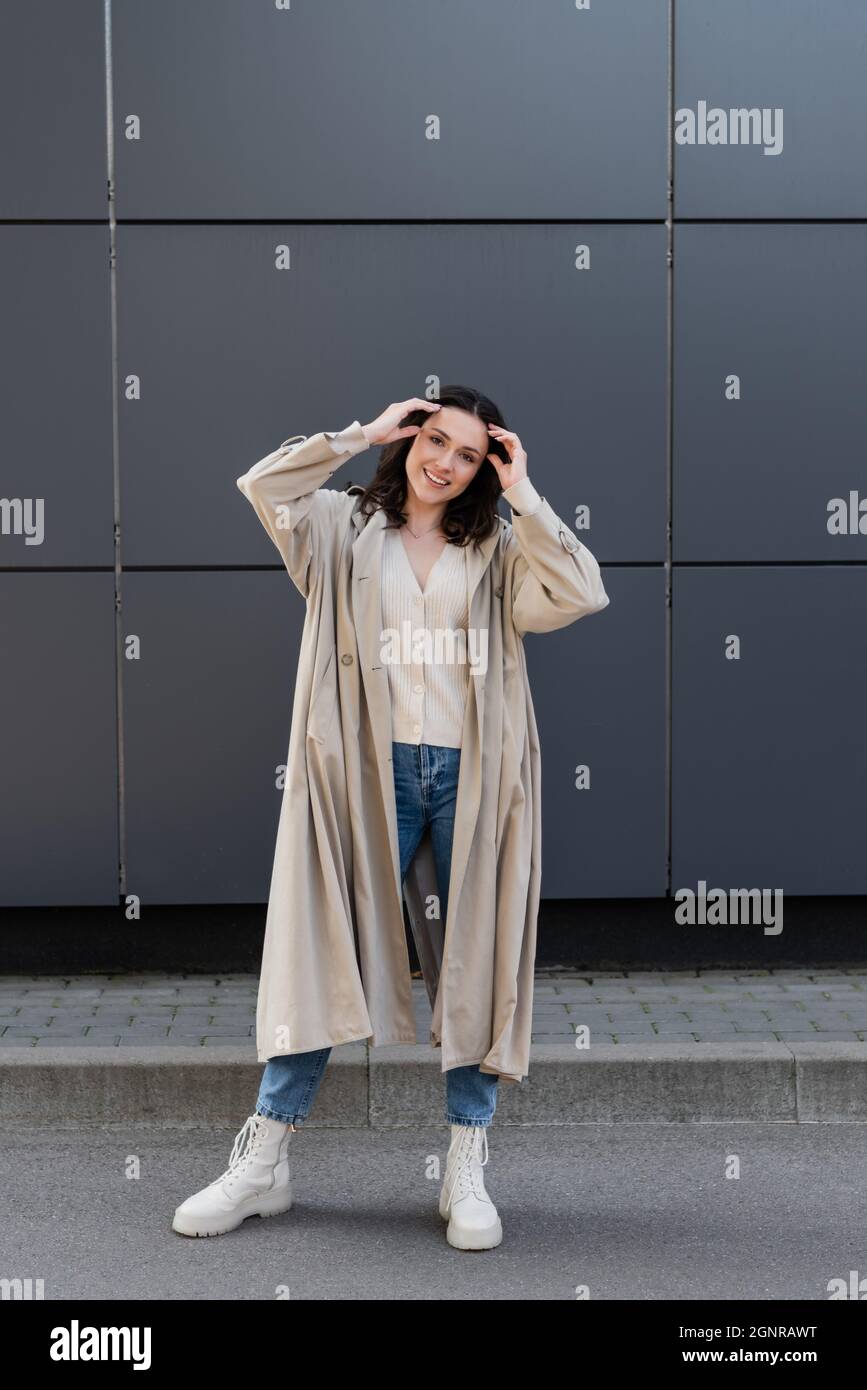 mujer sonriente con chubasquero largo y botas blancas que ajustan el pelo  cerca de la pared gris Fotografía de stock - Alamy