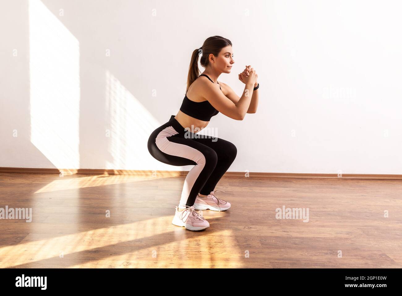 Retrato completo de una mujer haciendo ejercicio en sentadilla o en forma  de sentadilla en casa o gimnasio, con medias y camiseta deportiva negra.  Estudio interior iluminado por la luz del sol