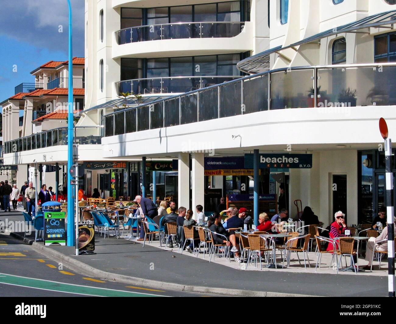 Los comensales se sientan en cafés en la acera a lo largo de Marine Parade tomando el sol de agosto en Mount Maunganui, Bay of Plenty, North Island, Nueva Zelanda. La tranquila ciudad al lado del océano es conocida por su volcán extinto, la playa del océano Pacífico y los cafés en la acera. Foto de stock