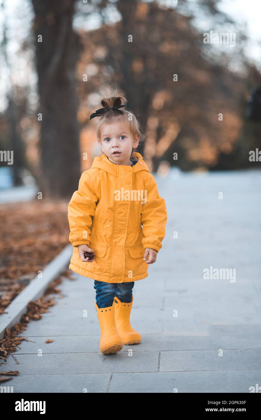 Bastante divertido niño niña de 2-3 años de edad usar chubasquero brillante, botas de goma en el parque sobre hojas caídas al aire libre. Temporada otoño. Feliz niño