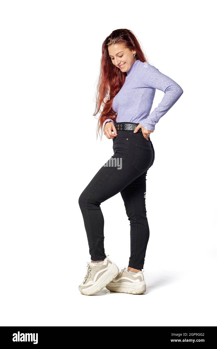 mujer joven en ropa informal posando y tocando el citurón sonriendo, con pelo rojo y ropa juvenil e informal Fotografía de stock -