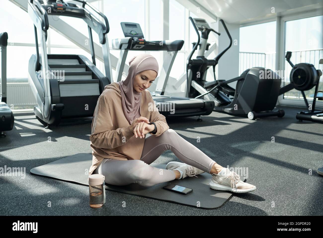 Disparo horizontal de una mujer musulmana moderna con hijab sentado en la colchoneta en el gimnasio que ajusta el dispositivo de seguimiento de actividad durante el tiempo de descanso Foto de stock