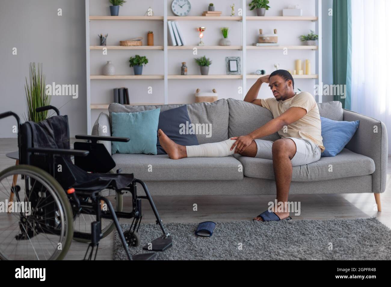 Un hombre negro infeliz que había vendado la pierna rota, sentado en el sofá, sufriendo de dolor en casa, silla de ruedas de pie cerca Foto de stock