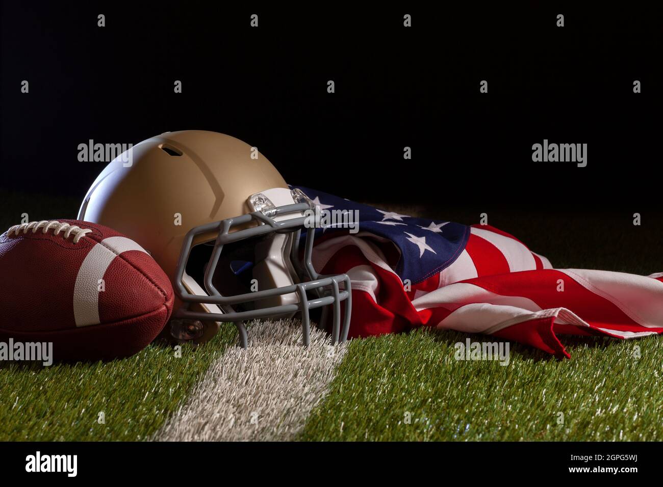 Vista en ángulo bajo de un balón de fútbol, casco y bandera estadounidense en un campo de hierba con rayas y fondo oscuro Foto de stock