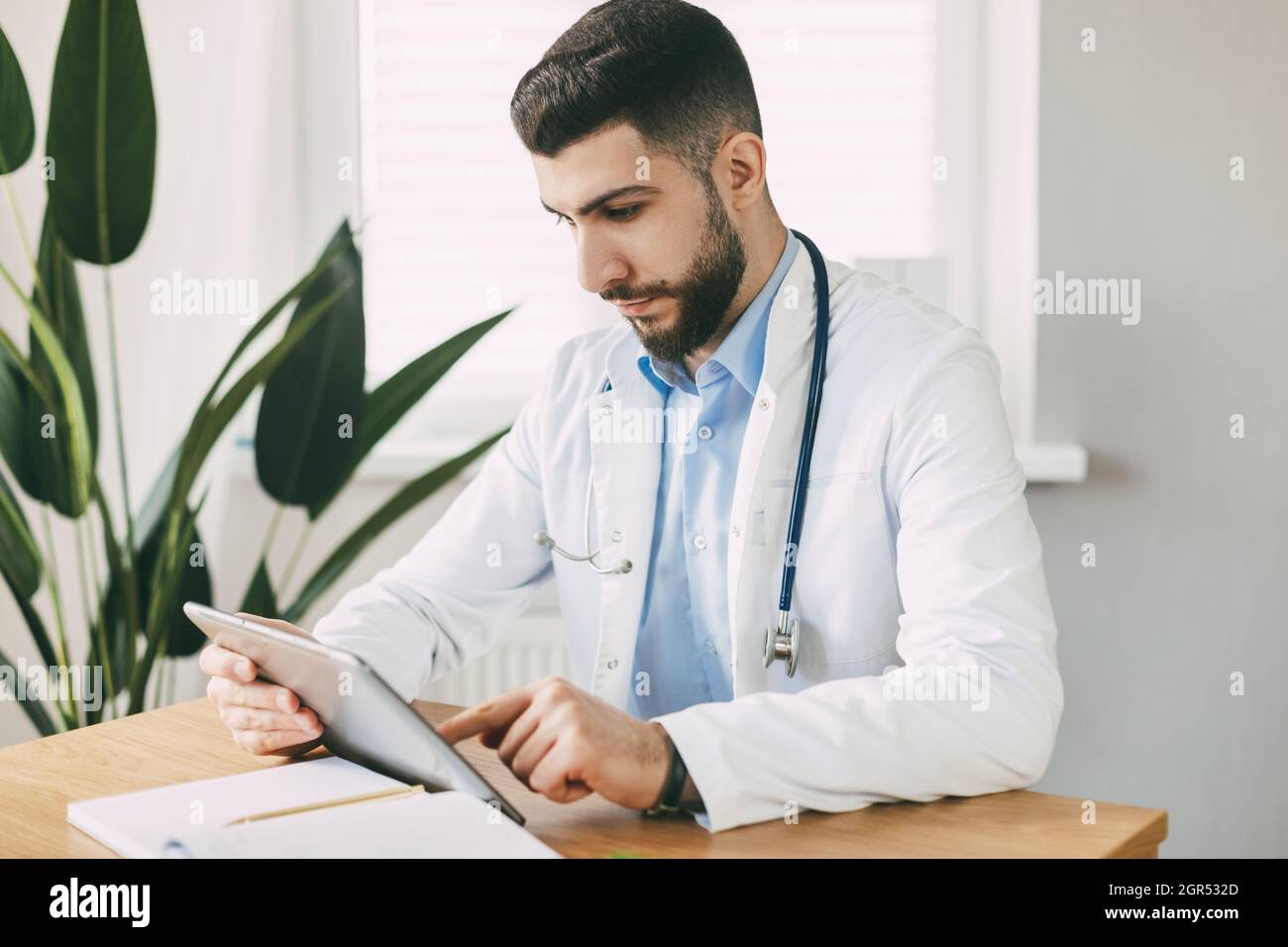 Un Doctor Masculino de Apariencia Oriental está Concentratedly trabajando en una Tableta, sentado en una Mesa Foto de stock
