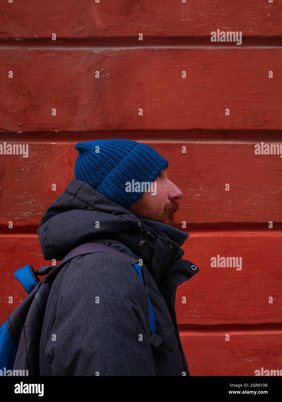 Chaqueta de senderismo con barba Guy en Blue Hat y abrigo gris con mochila  sobre fondo de pared rojo Fotografía de stock - Alamy