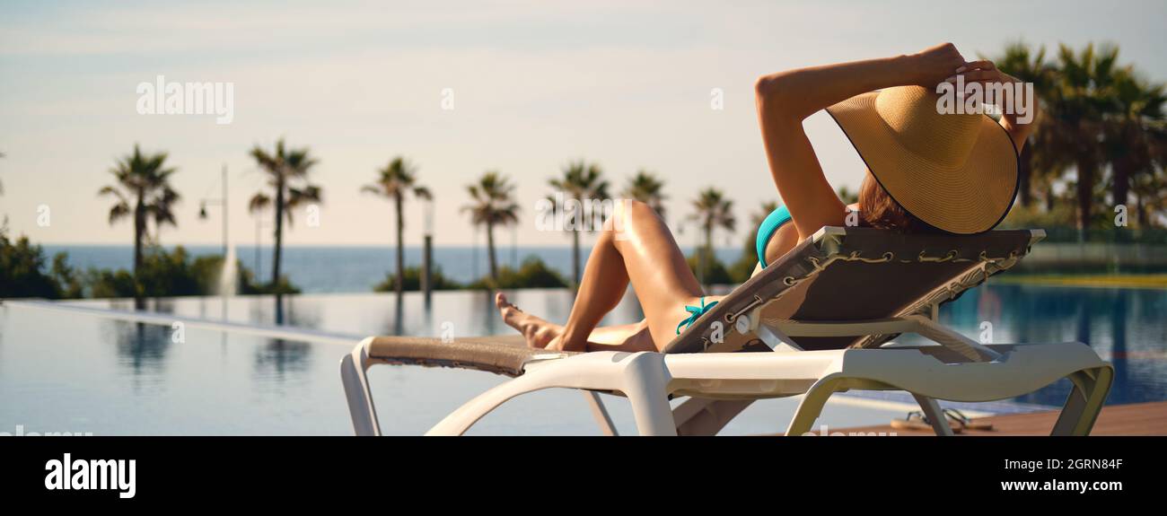 Vista trasera mujer llevar sombrero tumbado en tumbona cerca de la piscina, poner las manos detrás de la cabeza relajante, tomar el sol baño, paisaje tropical en el fondo. Vacaciones de verano Foto de stock
