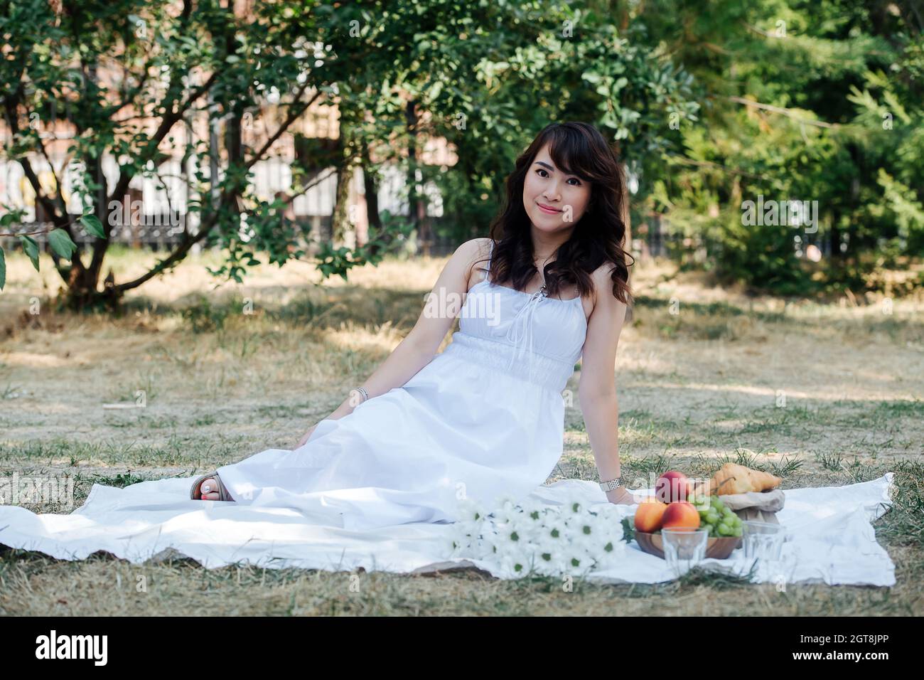 Retrato elegante de una encantadora mujer asiática elevada sentada sobre  una manta en un parque. Ella lleva vestido blanco, haciendo picnic en un  parque, disfrutando de la última wa Fotografía de stock -