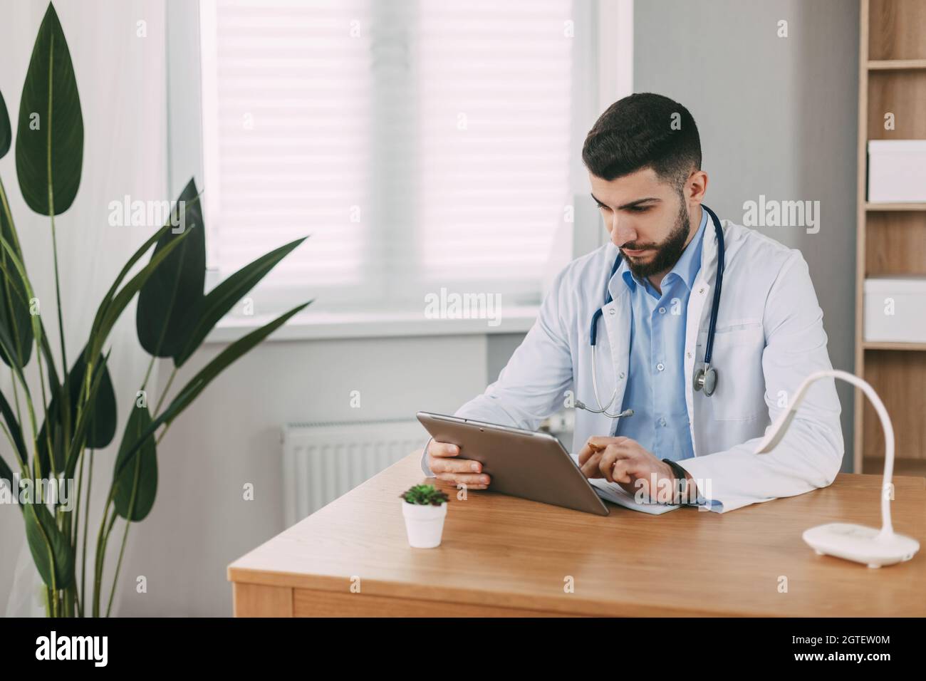 Un Doctor Masculino de Apariencia Oriental está Concentratedly trabajando en una Tableta, sentado en una Mesa Foto de stock