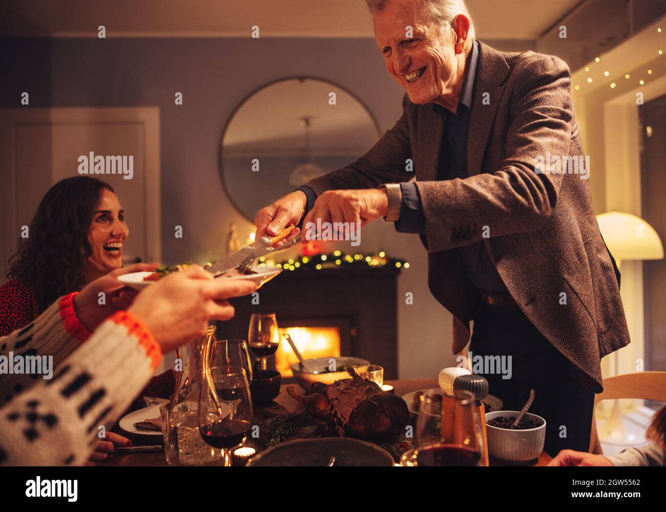 Hombre mayor que sirve comida a la familia sentado en la mesa de la cena. Cena de Navidad en casa escandinava. Foto de stock