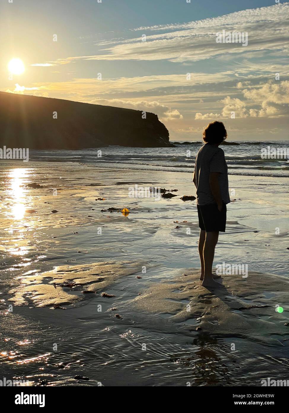 Vista trasera completa de un hombre maduro de pie en la playa a última hora de la tarde cuando se pone el sol. Mirando hacia el horizonte usando pantalones cortos y camiseta. Foto de stock