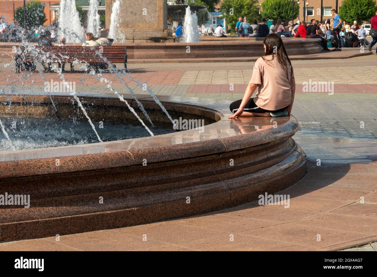 Una joven se sienta sola al borde de una fuente en la plaza de la estación de tren llena de gente en un día caluroso de verano. Foto de stock