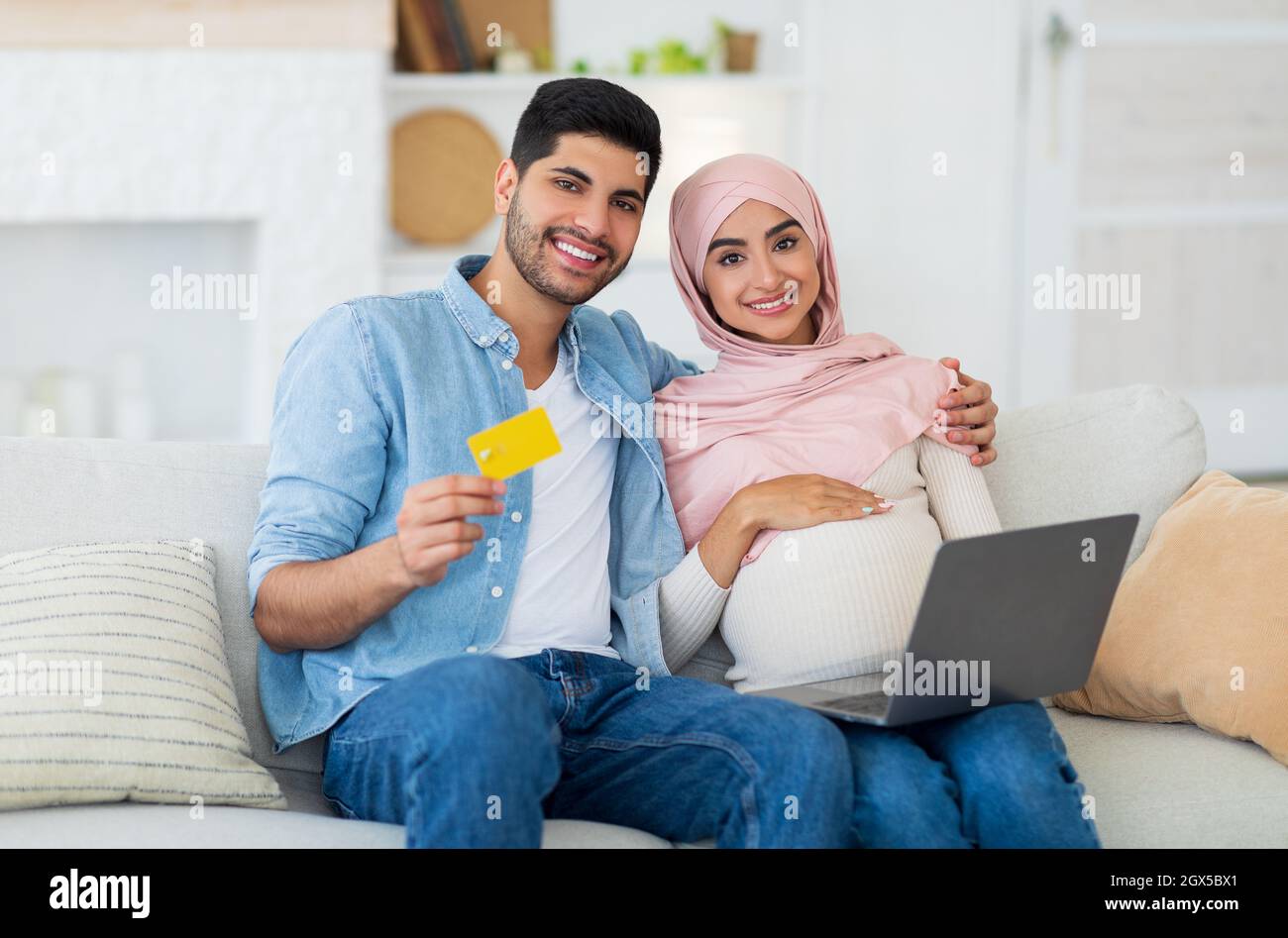 Felices cónyuges islámicos embarazadas disfrutando de hacer pagos en línea con el ordenador portátil y la tarjeta de crédito, descansando en casa Foto de stock