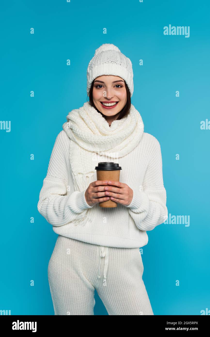 Mujer bonita en bufanda de punto y sombrero sosteniendo café para ir aislado en azul Foto de stock