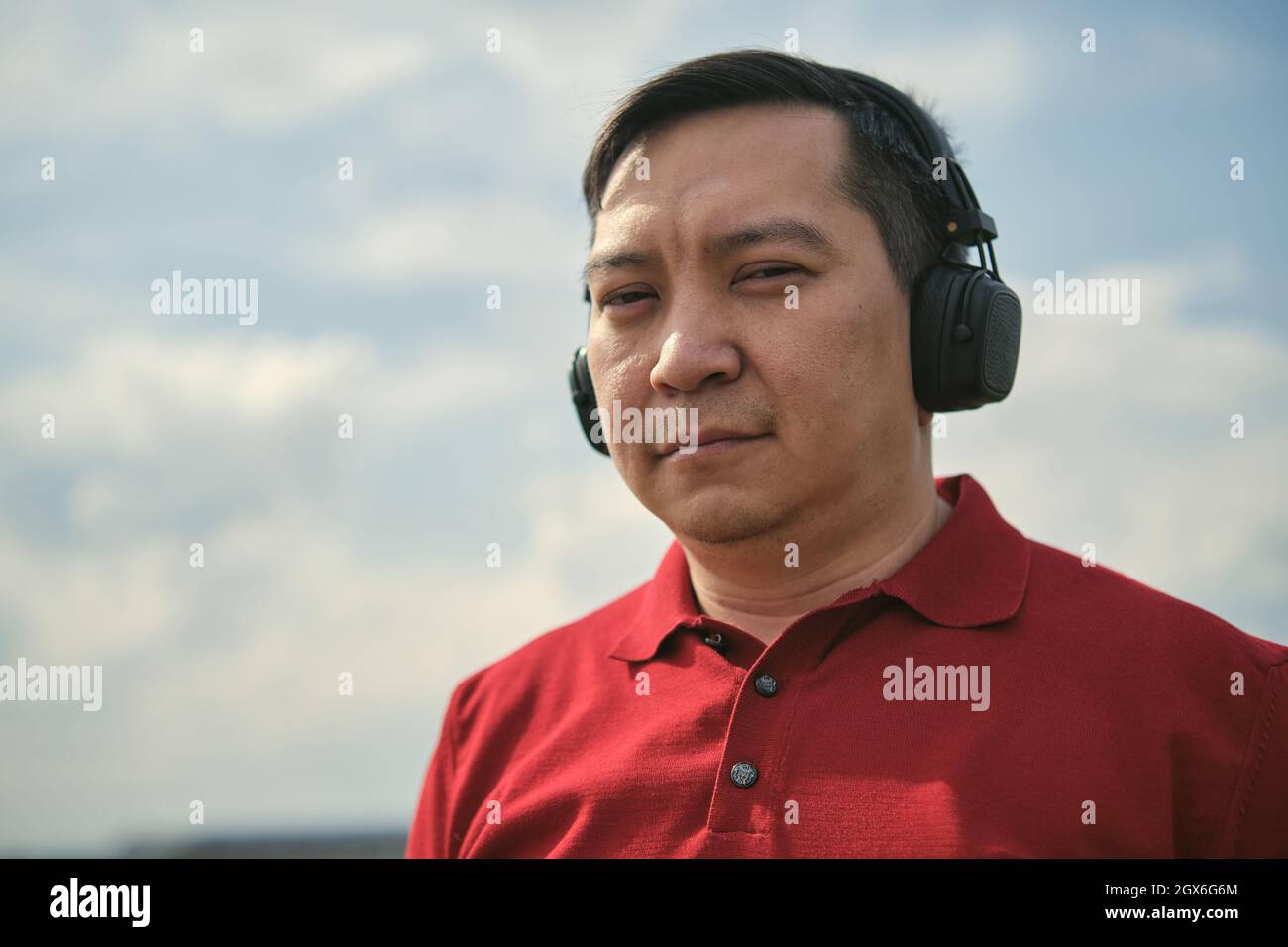 Hombre asiático de mediana edad con auriculares al aire libre escuchando música sobre el fondo del cielo Foto de stock