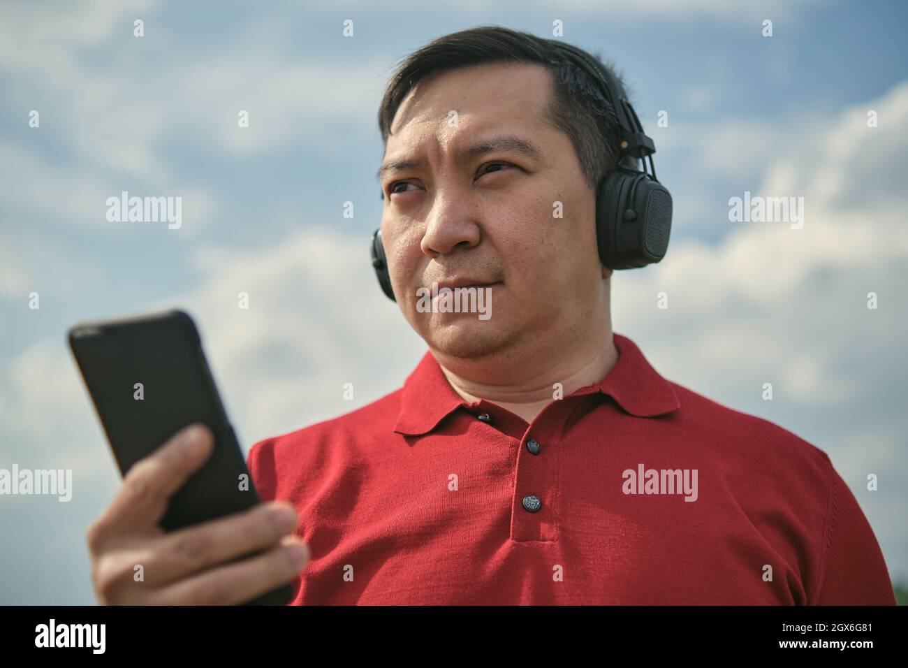 Hombre asiático de mediana edad con auriculares al aire libre escuchando música sobre el fondo del cielo, teléfono móvil en la mano Foto de stock