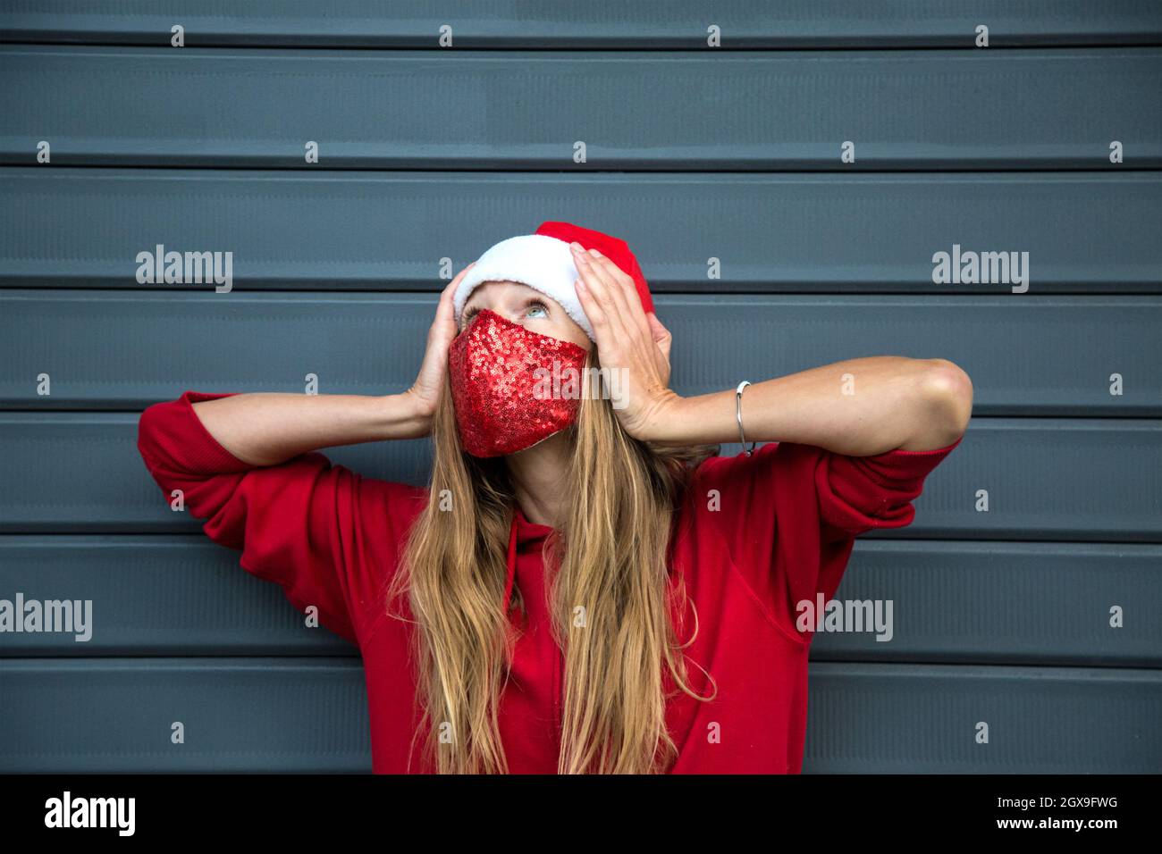 Longhair rubia caucásica chica en rojo sombrero de Navidad, sudadera con capucha y máscara protectora brillante sostiene la cabeza, mira hacia arriba. No sé qué hacer gesto Foto de stock