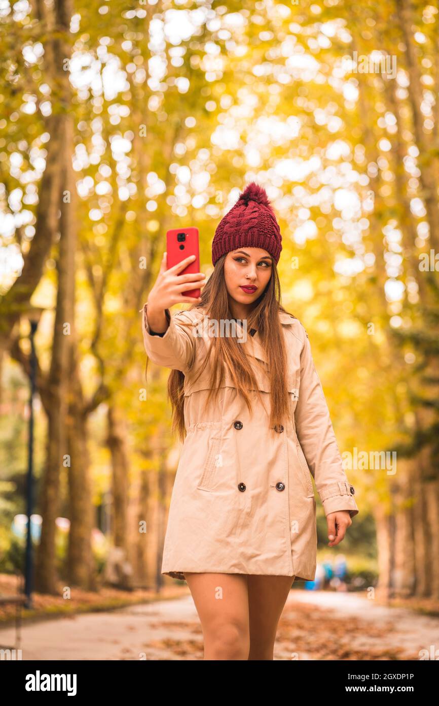Joven mujer con estilo en abrigo de trinchera y sombrero de punto que se autorretrato en el teléfono móvil en callejón entre los árboles en la temporada de otoño Foto de stock