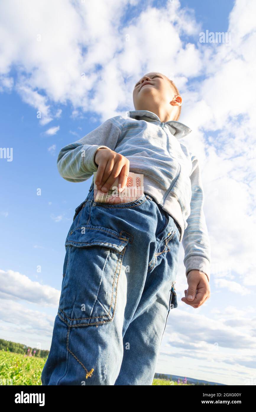 Un niño de preescolar satisfecho en jeans y un suéter sostiene el dinero  del papel en la naturaleza en un campo contra el fondo de un cielo azul con  nubes. Vertical Fotografía