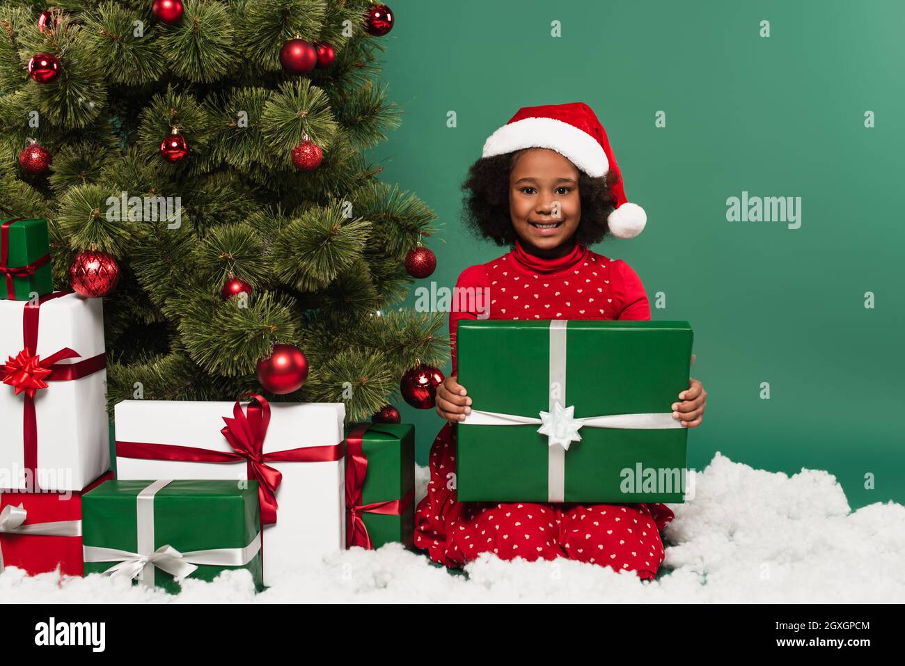 Niño afroamericano en sombrero de santa con regalo cerca del árbol de  Navidad y nieve decorativa sobre fondo verde Fotografía de stock - Alamy