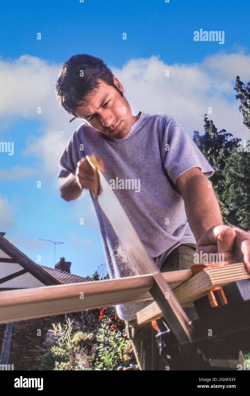 DIY Handyman casa de mejora de mantenimiento 30-40 años en el hogar DIY handyman que trabaja al aire libre en el jardín de la casa, aserrando una tabla de madera en un banco de trabajo, ángulo bajo fuera con el cielo azul de la casa y las nubes en el fondo Foto de stock
